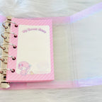 A pink ring-bound planner or notepad set shown open on a white faux-fur surface. The cover has diagonal pink stripes, a blank writing area, and a cartoon character near the bottom.