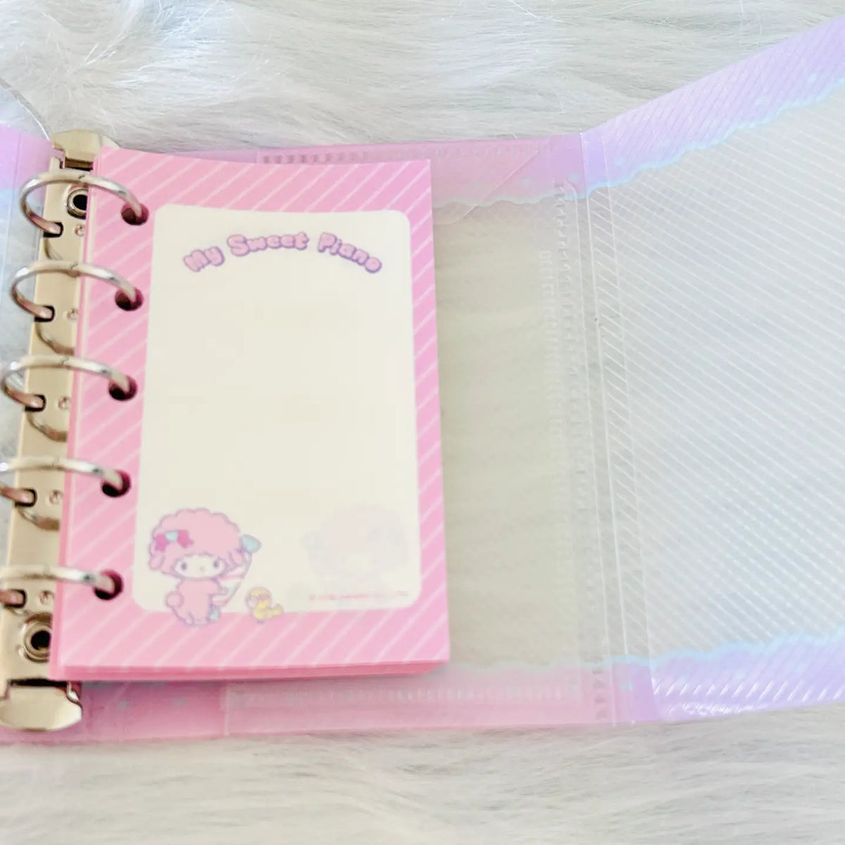 A pink ring-bound planner or notepad set shown open on a white faux-fur surface. The cover has diagonal pink stripes, a blank writing area, and a cartoon character near the bottom.