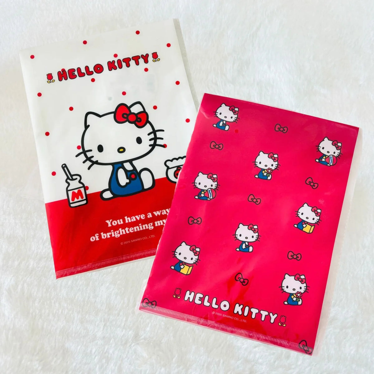Two Hello Kitty plastic file folders on a white textured fabric background. One folder is white with red polka dots, a red bottom band, and a large Hello Kitty illustration.