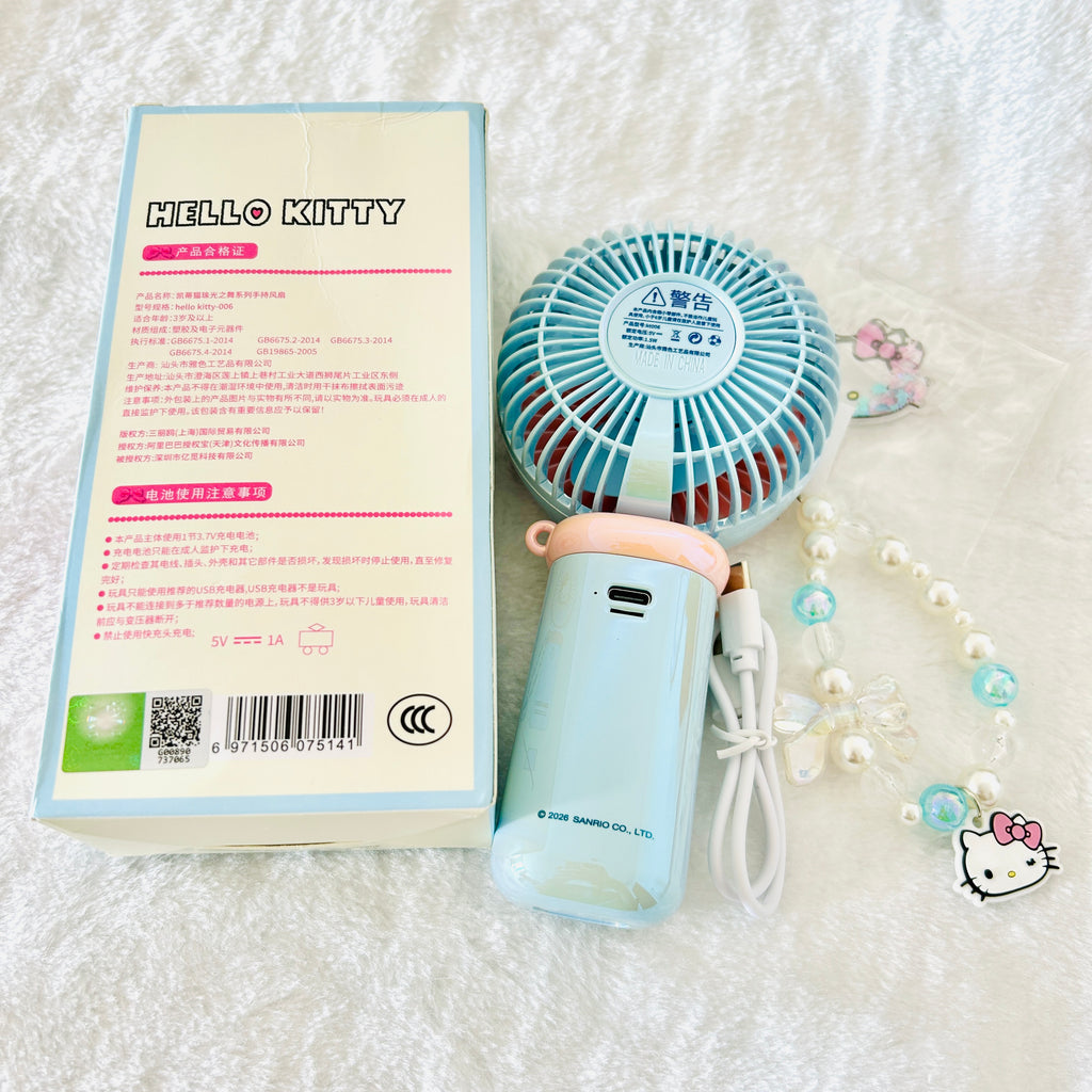 A light blue and pink Hello Kitty portable fan shown next to its product packaging box and a decorative Hello Kitty beaded necklace on a white textured fabric background.