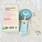 A light blue and pink Hello Kitty portable fan shown next to its product packaging box and a decorative Hello Kitty beaded necklace on a white textured fabric background.