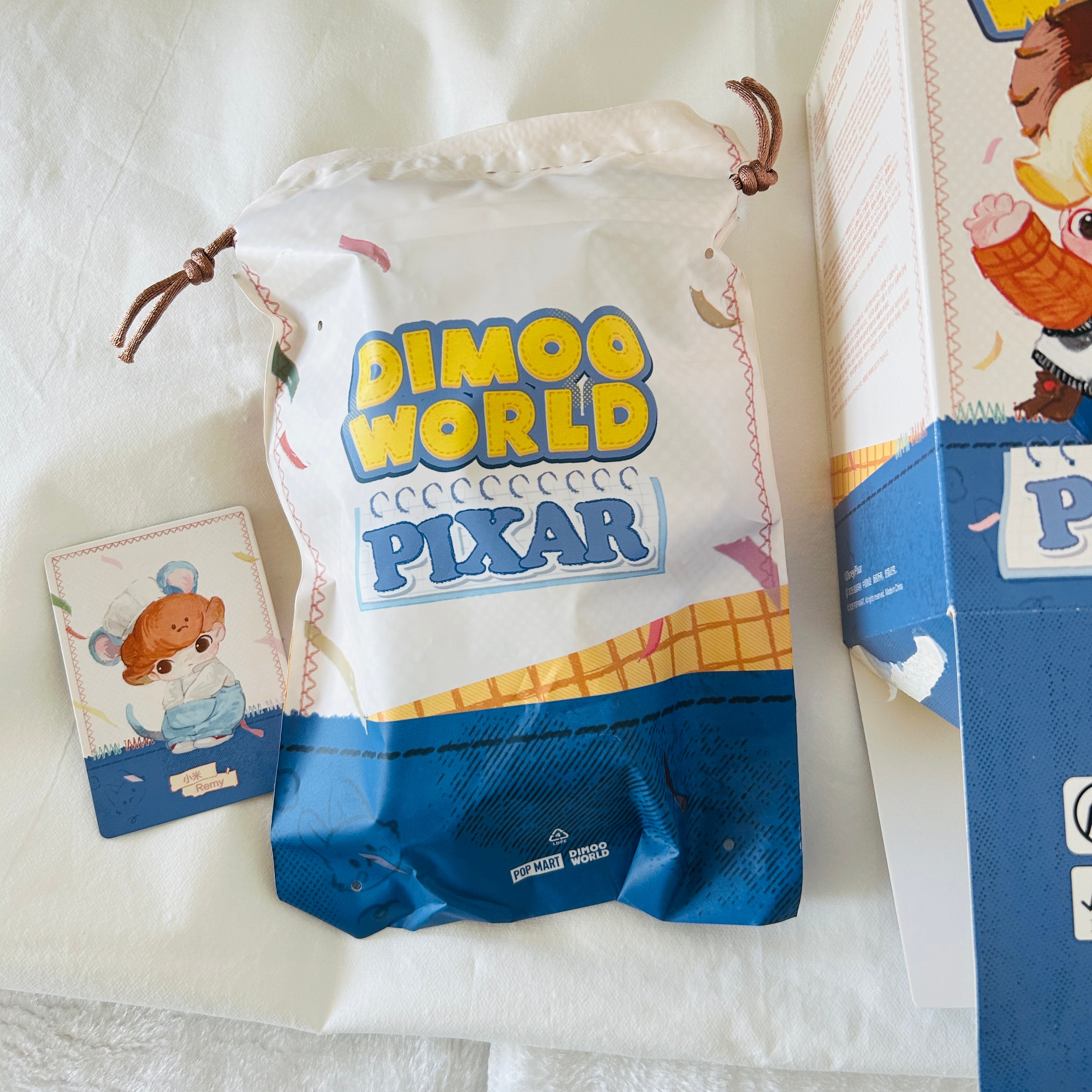 A Dimoo World Pixar branded drawstring pouch with a blue lower section and cartoon character art, shown Remy is the character.