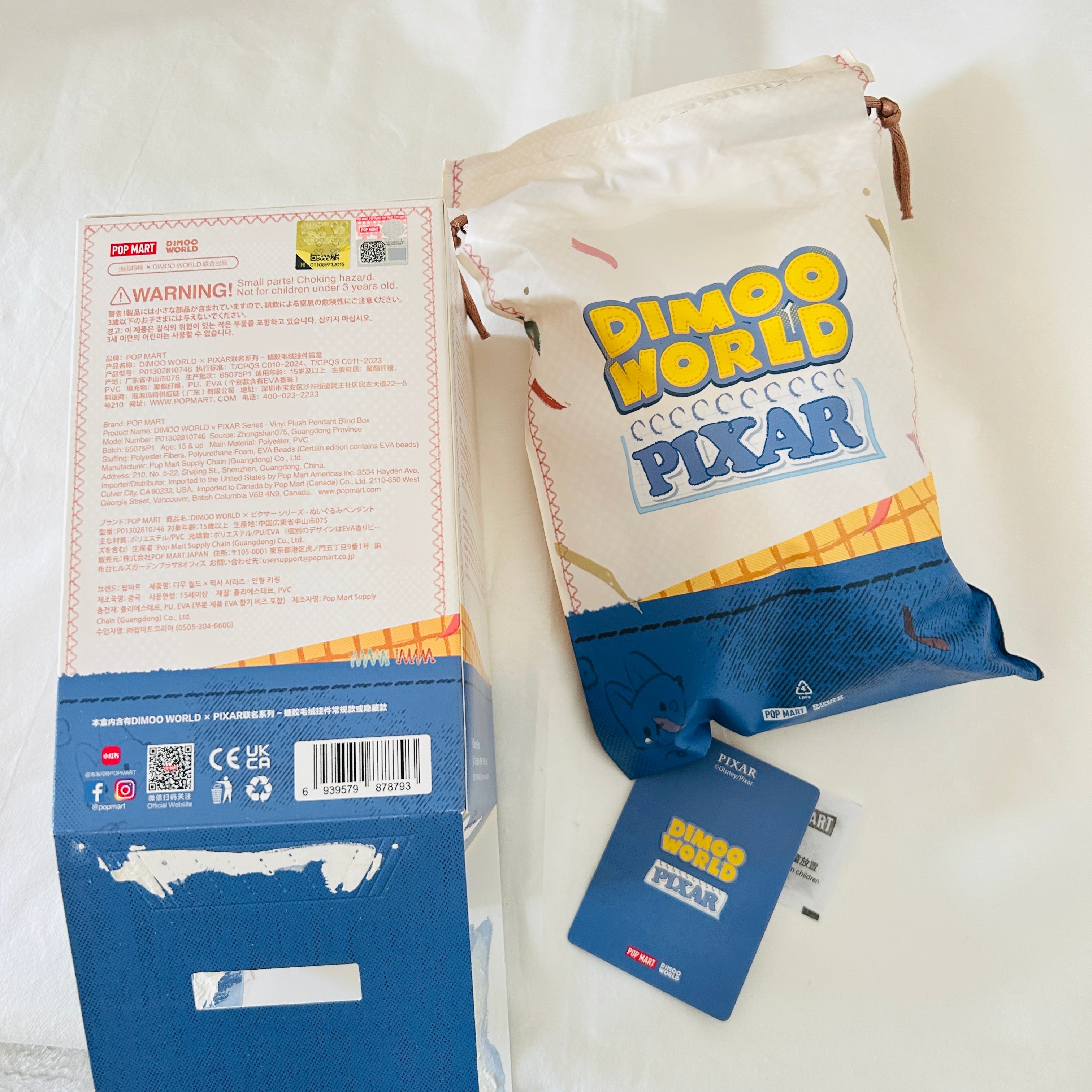 A sealed Dimoo World Pixar collectible pouch lies on a white surface next to its blue cardboard packaging box and a blue hangtag. The pouch is off-white with a blue lower section and large yellow and blue 'Dimoo World Pixar' text on the front.
