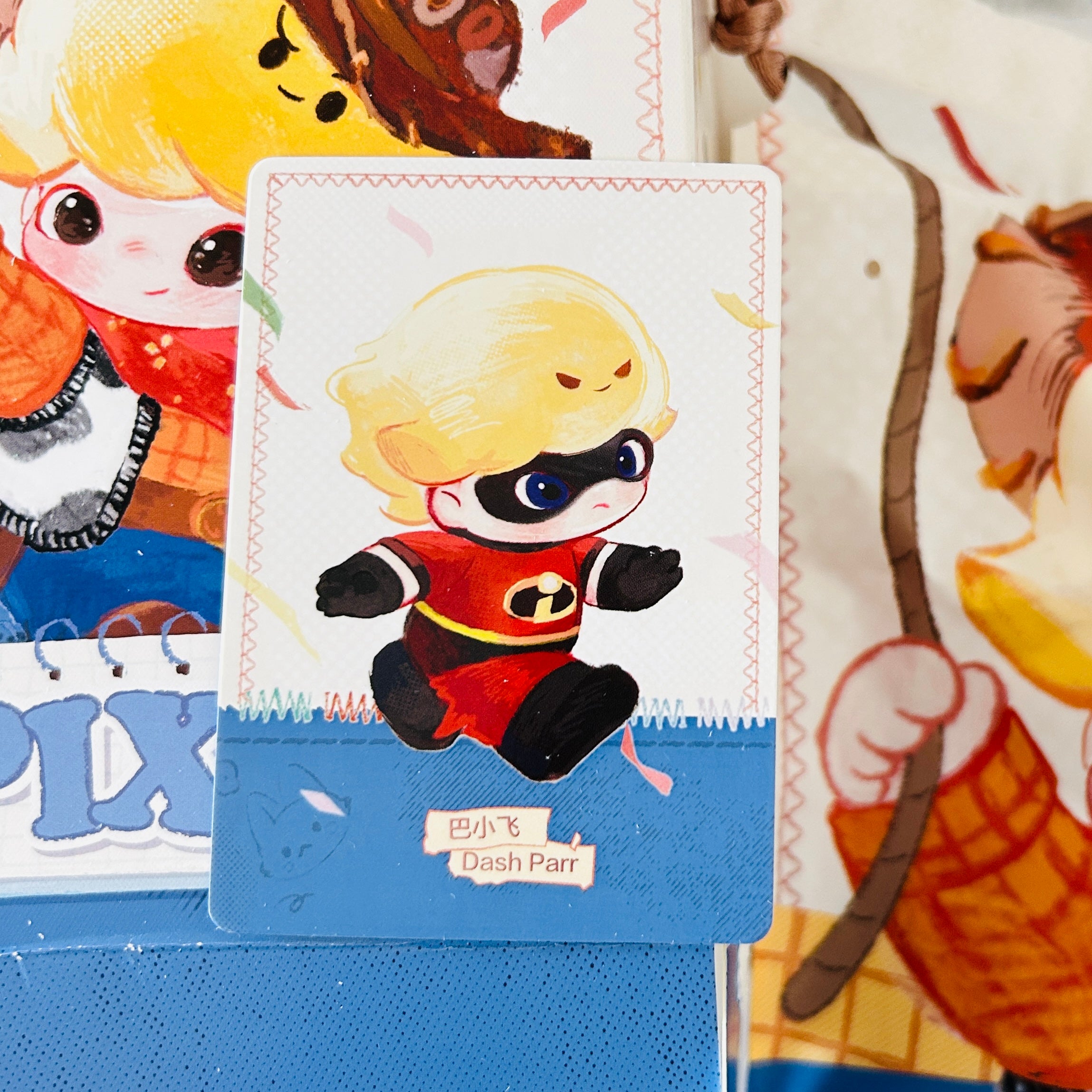 A close-up of a collectible card featuring a chibi-style cartoon character inspired by Dash from The Incredibles, with Chinese text reading 'Dash Parr' on a blue base. Other illustrated cards are partially visible in the background.