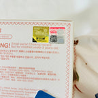 Close-up of the back of a product box with printed text in English and Japanese, warning labels, a QR code, and a small gold/yellow product photo near the top. A blue object is partially visible at the bottom edge.