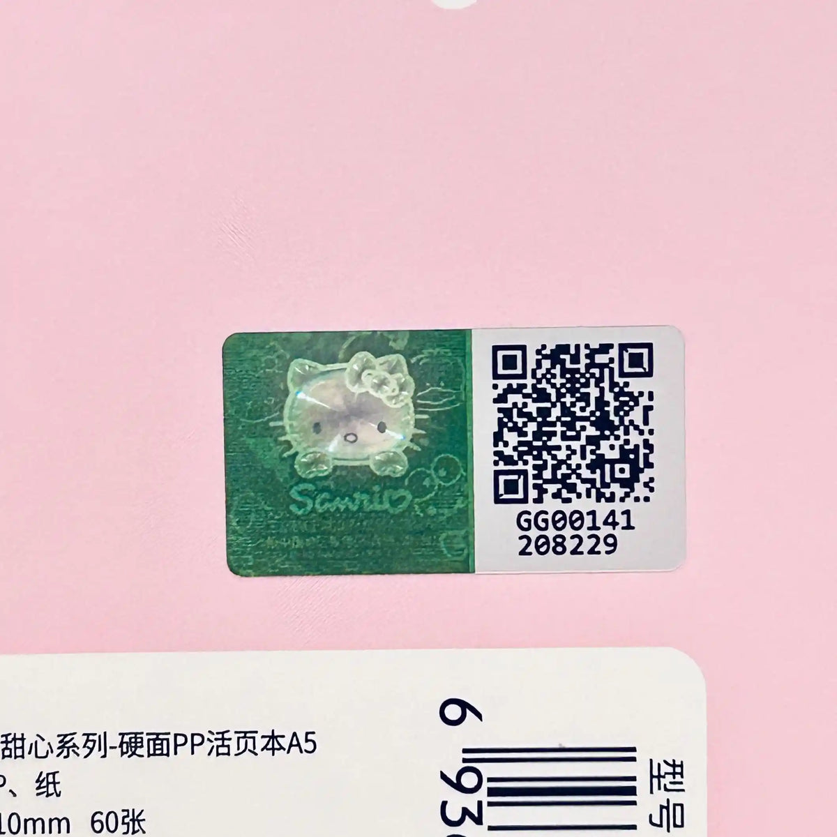A close-up of a pink product surface showing a rectangular Sanrio sticker with a green Hello Kitty design and a QR code, plus part of a white barcode label below.