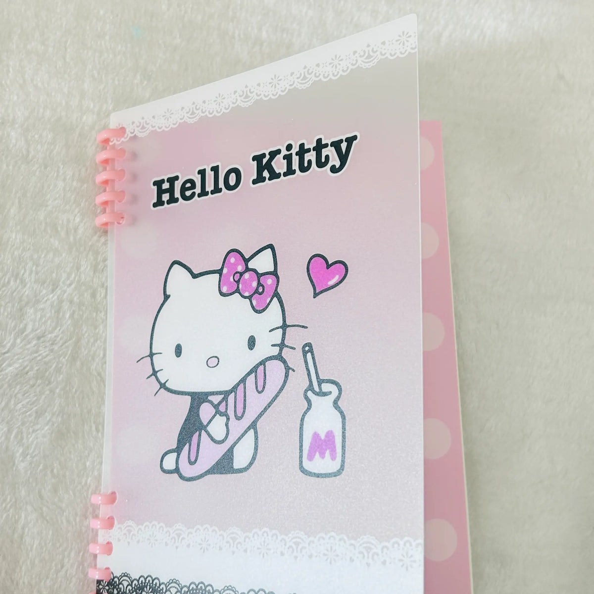 A pink Hello Kitty spiral-bound notebook or planner cover featuring the Hello Kitty character, a pink bow, and a milk bottle illustration on a soft white textured background.