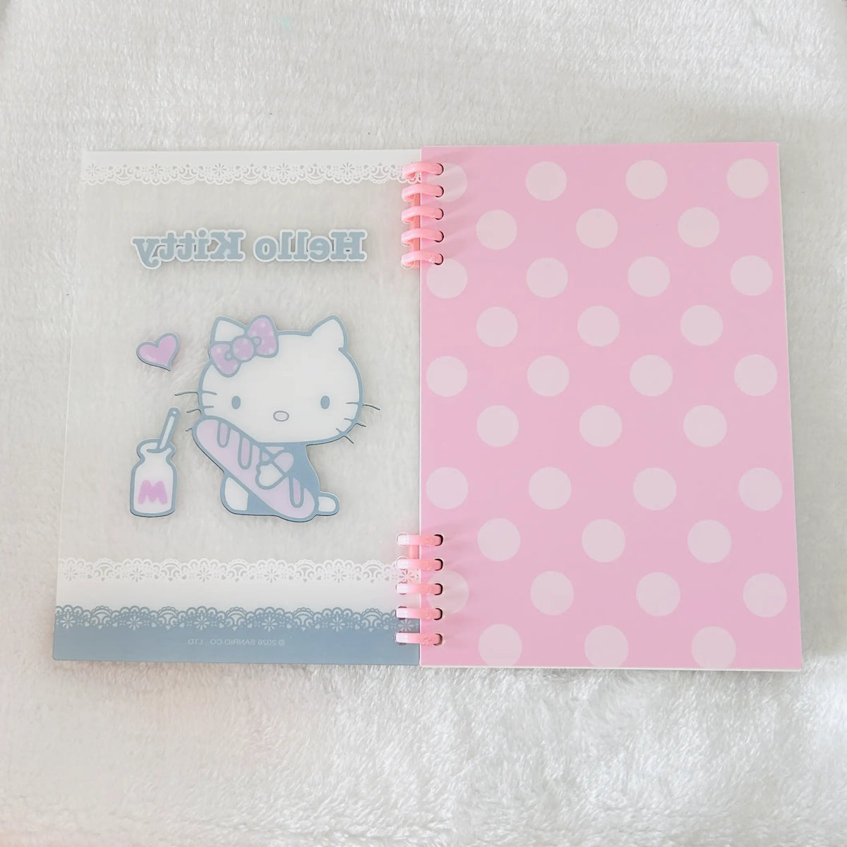 A pink and white Hello Kitty spiral-bound notebook. The left page has a Hello Kitty illustration with the text 'Hello Kitty' and a small milk bottle graphic, while the right page is pink with white polka dots.