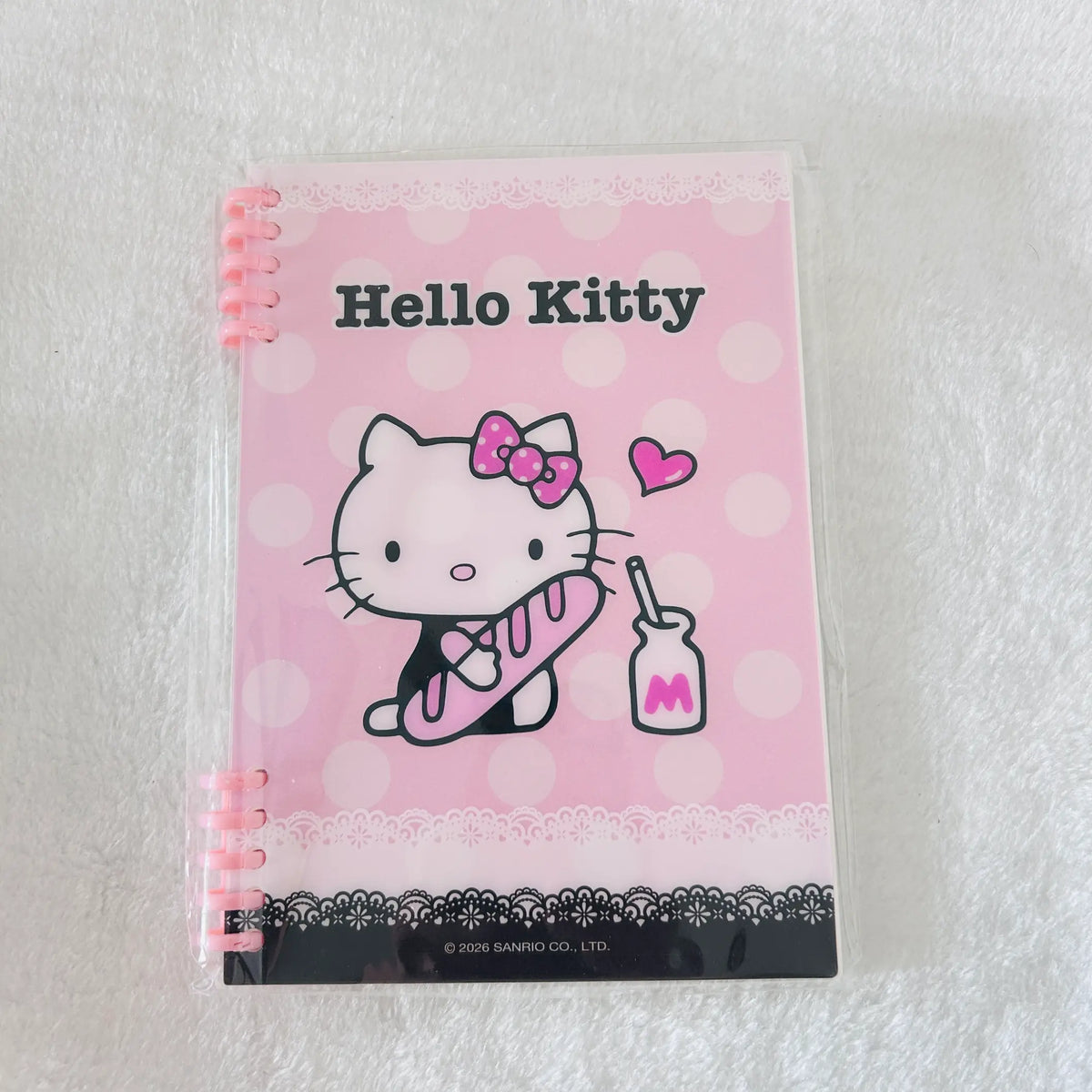 A pink Hello Kitty spiral notebook in a clear plastic sleeve, resting on a white textured fabric background. The cover features the Hello Kitty character holding a pink lollipop, with 'Hello Kitty' printed at the top.