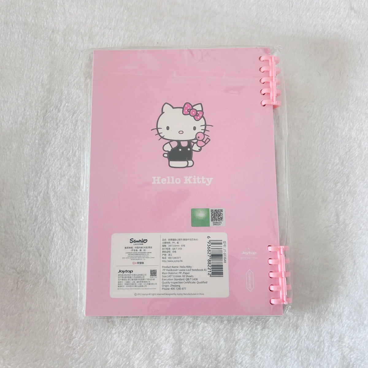 A pink Hello Kitty spiral notebook in its clear plastic protective sleeve, placed on a white textured fabric background. The notebook features the classic Hello Kitty illustration and Sanrio branding.