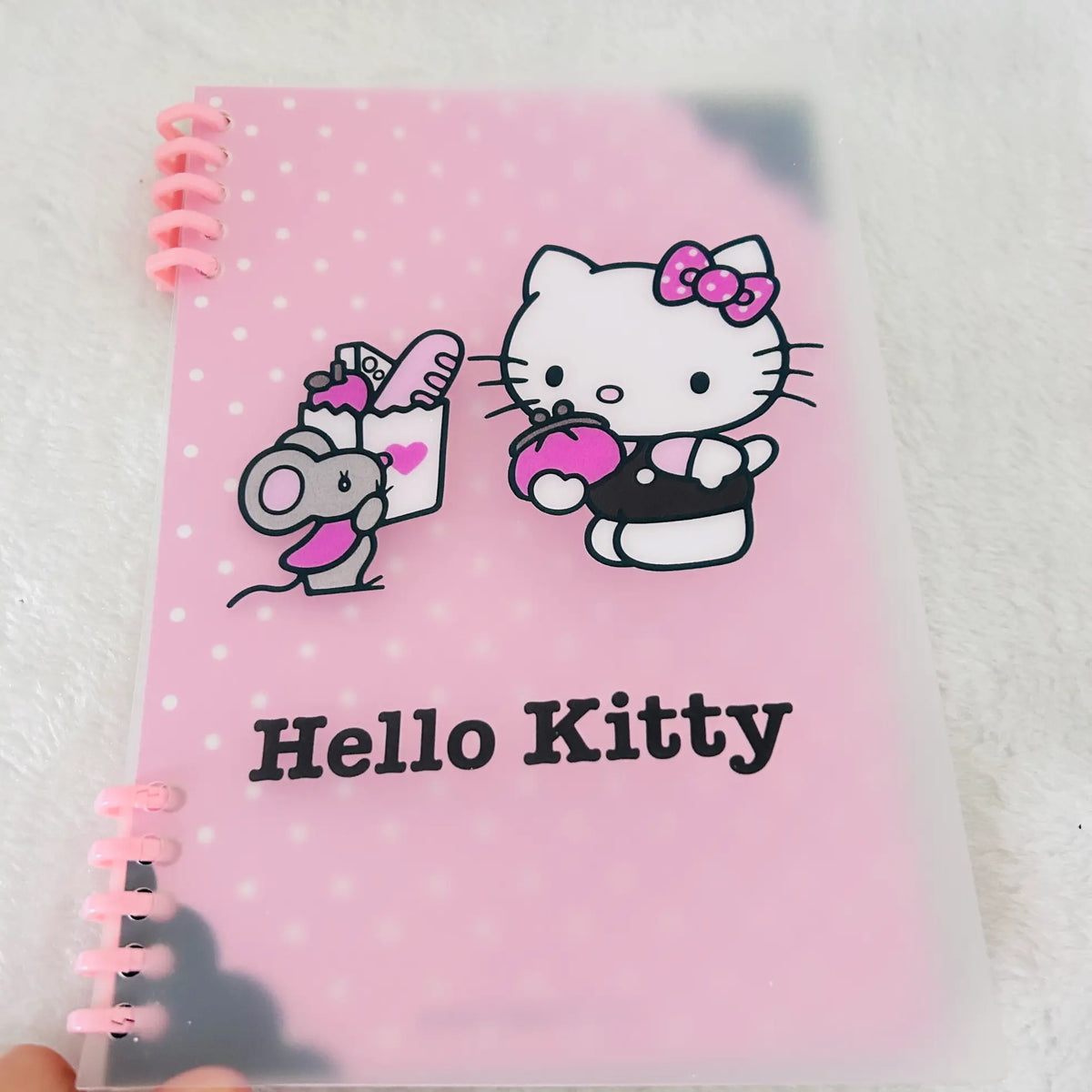 A pink Hello Kitty spiral notebook in a clear plastic sleeve. The cover features the Hello Kitty character with a mice holding a bag of food.