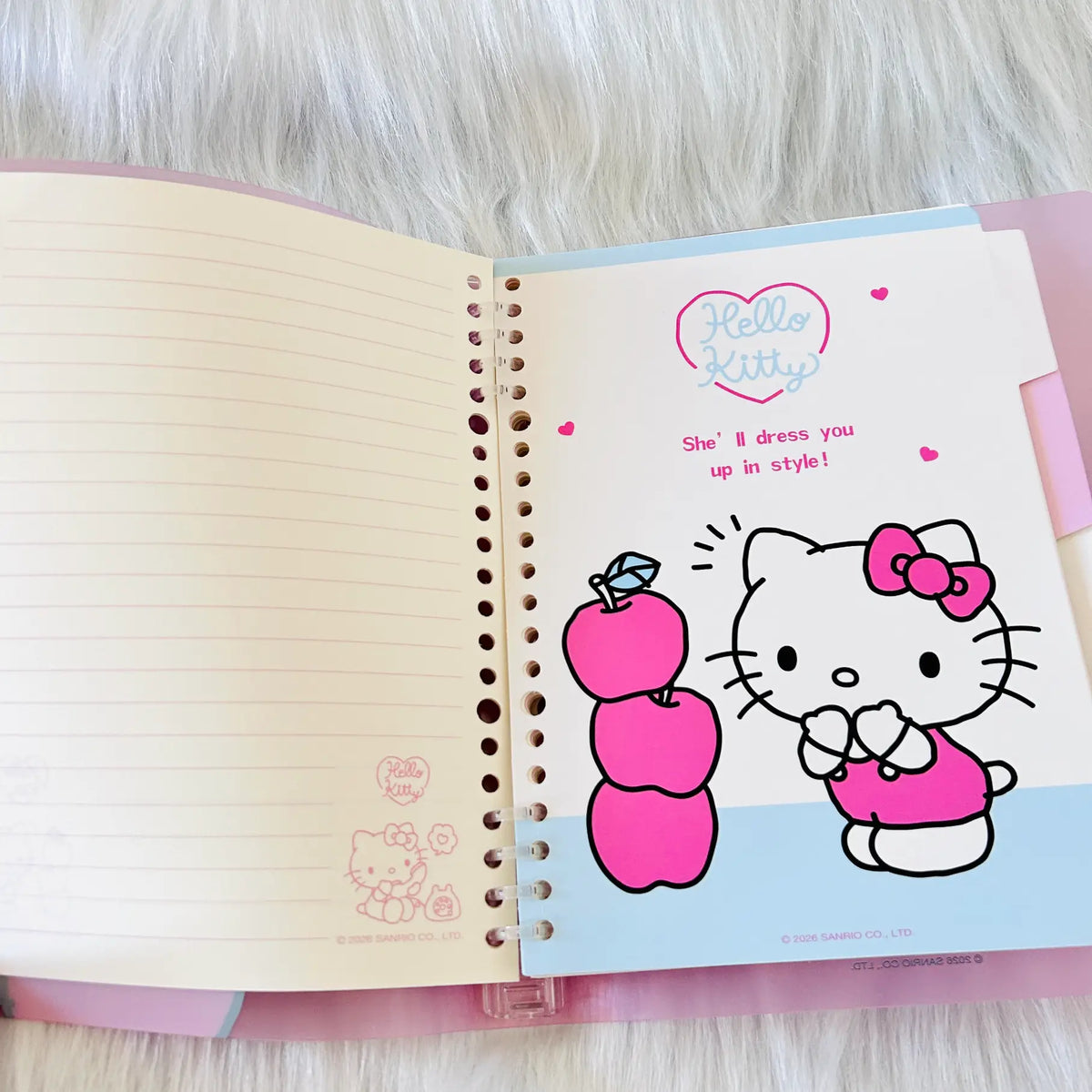 An open Hello Kitty spiral notebook  shows a page of divider with coloured Hello Kitty illustration, red apples and pastel blue and pink borders.