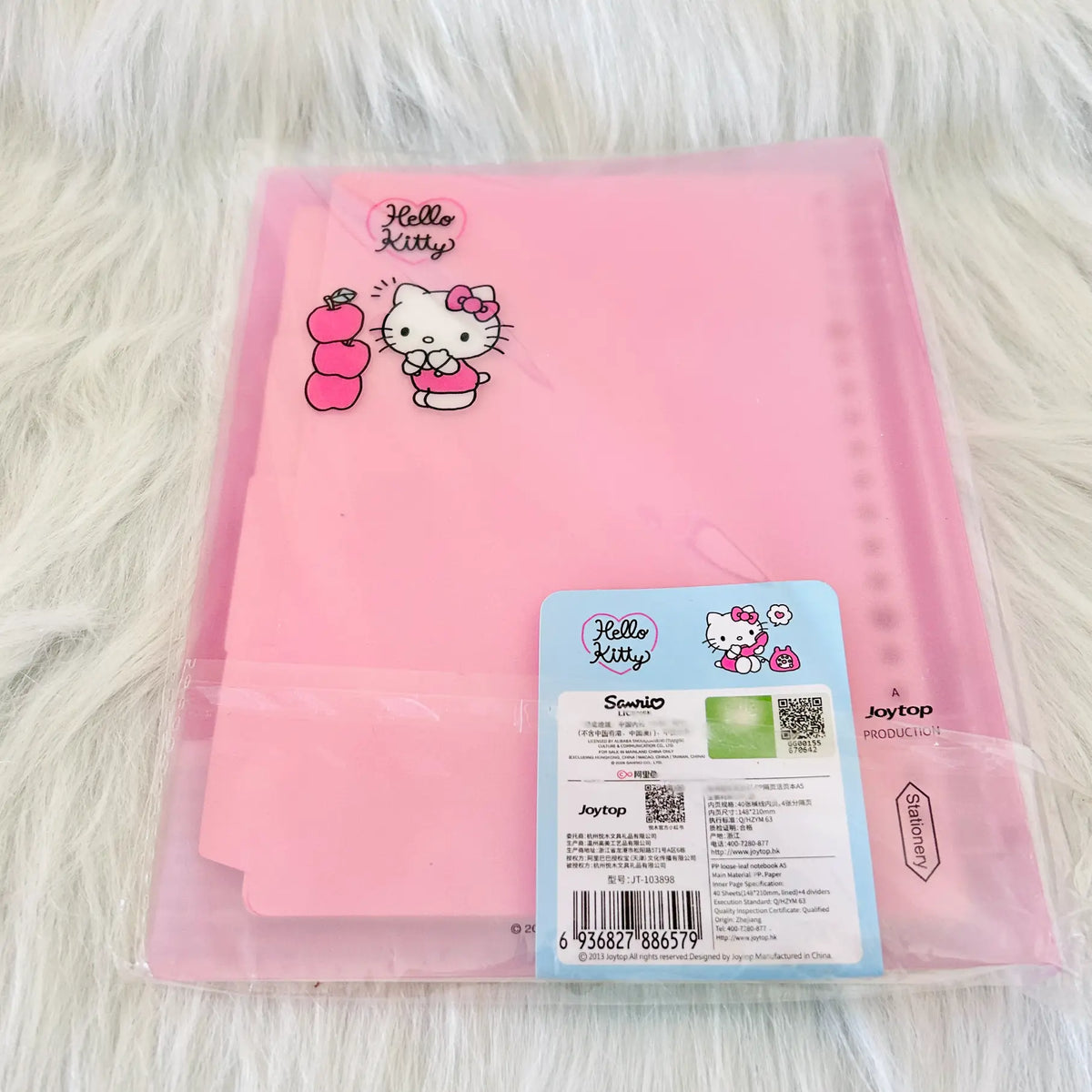 A pink Hello Kitty file folder inside a clear plastic sleeve, displayed on a white faux fur surface. The folder features a Hello Kitty character graphic and a pink apple illustration, with a blue and white product label attached at the bottom right.