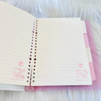 An open Hello Kitty spiral-bound lined notebook with cream-colored pages and pink illustrations, resting on a pink folder over a white faux fur background.