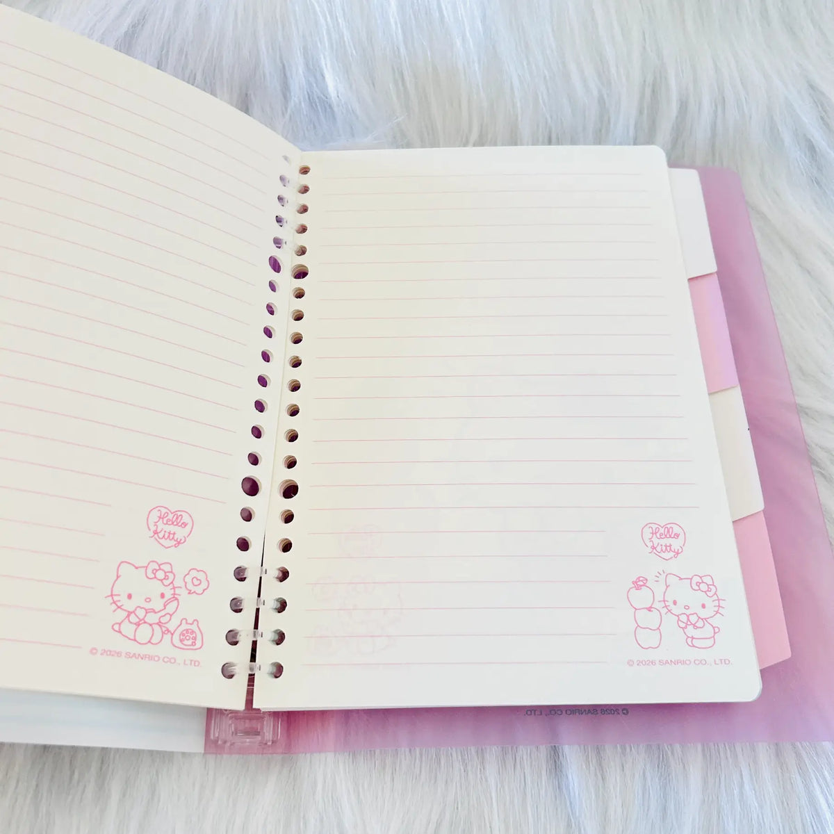 An open Hello Kitty spiral-bound lined notebook with cream-colored pages and pink illustrations, resting on a pink folder over a white faux fur background.