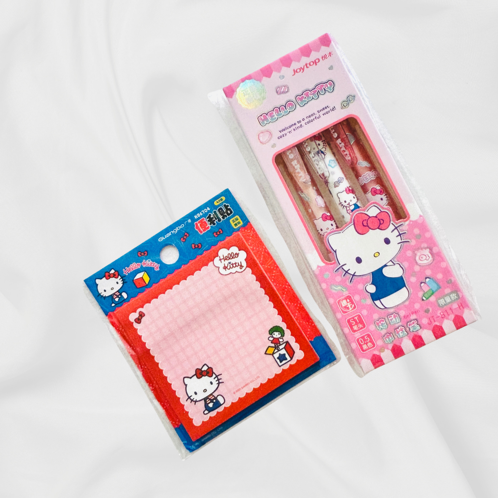 Hello Kitty stationery set including a pink memo pad in blue packaging and a pink pen set in a clear windowed box, both featuring Hello Kitty illustrations on a white fabric background.