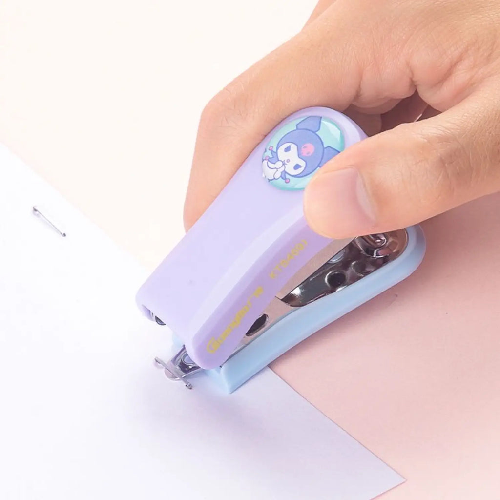 A hand holding a small pastel purple and light blue stapler with a cartoon character graphic on top, pressing it onto white paper.