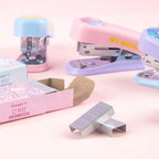 Three pastel-colored office staplers with matching paper boxes on a soft pink background, with loose silver metal staples in the foreground.