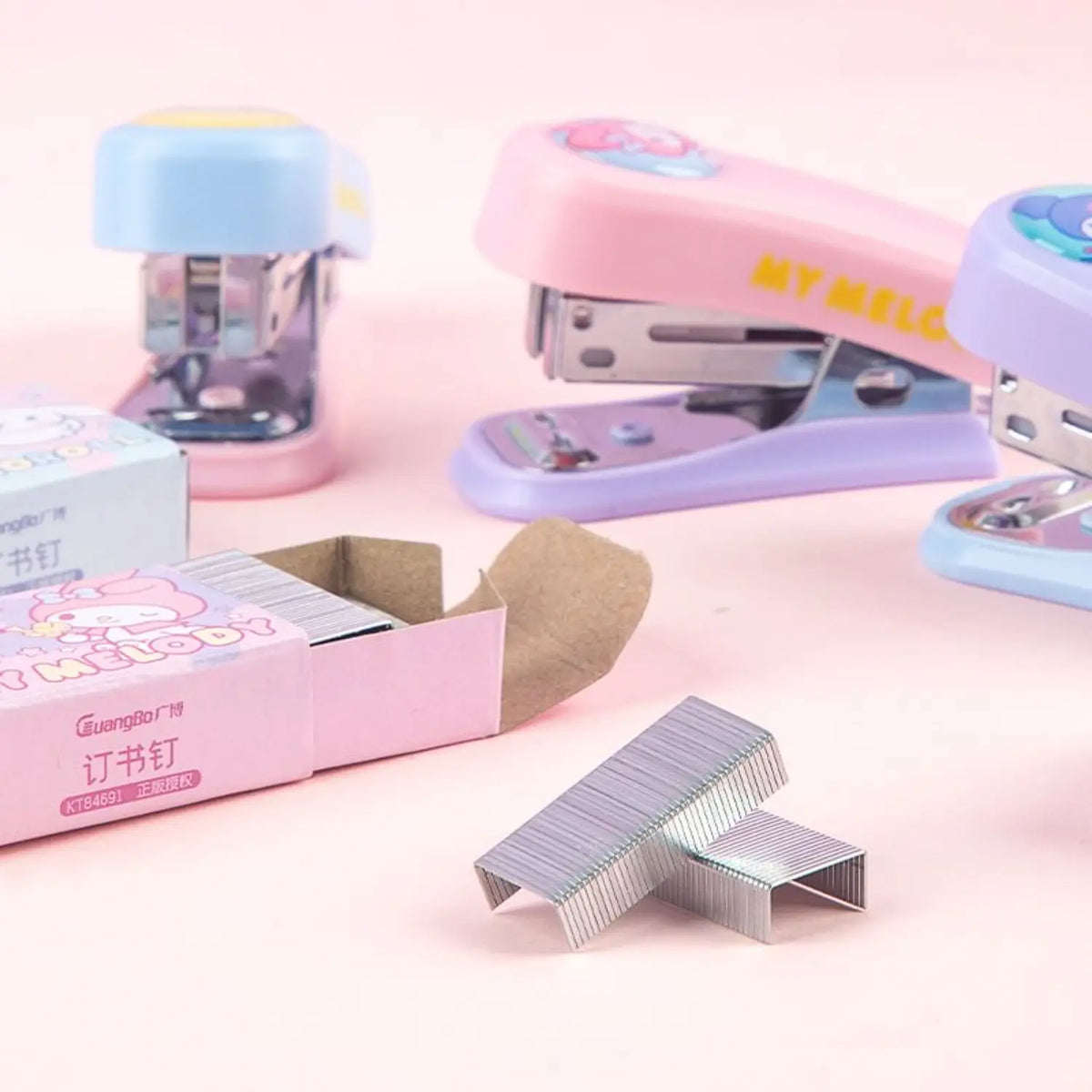 Three pastel-colored office staplers with matching paper boxes on a soft pink background, with loose silver metal staples in the foreground.