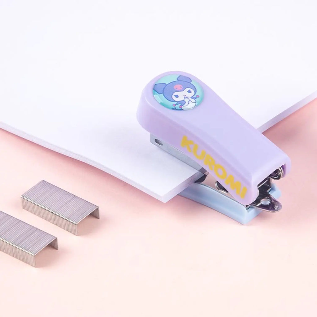 A small Kuromi-themed paper clip in lavender purple with yellow 'KUROMI' text and a cartoon character graphic, shown holding white paper. Two silver metal paper clips are placed beside it on a soft pink surface.