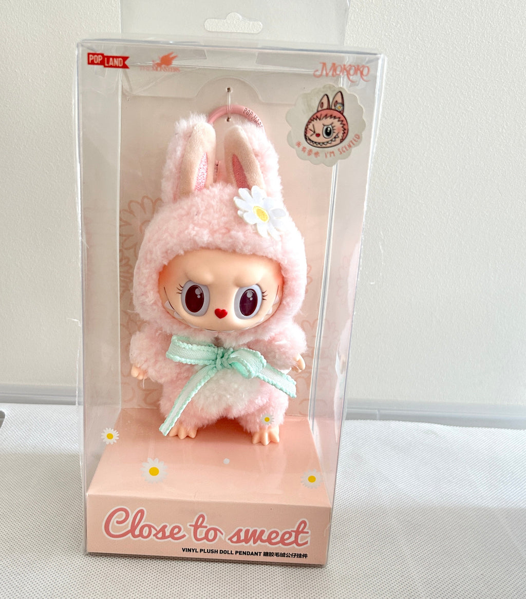 A plush doll character in a pink bunny outfit with a mint green bow, displayed inside a clear plastic retail box on a pink base. The box has 'Close to sweet' printed on the front.