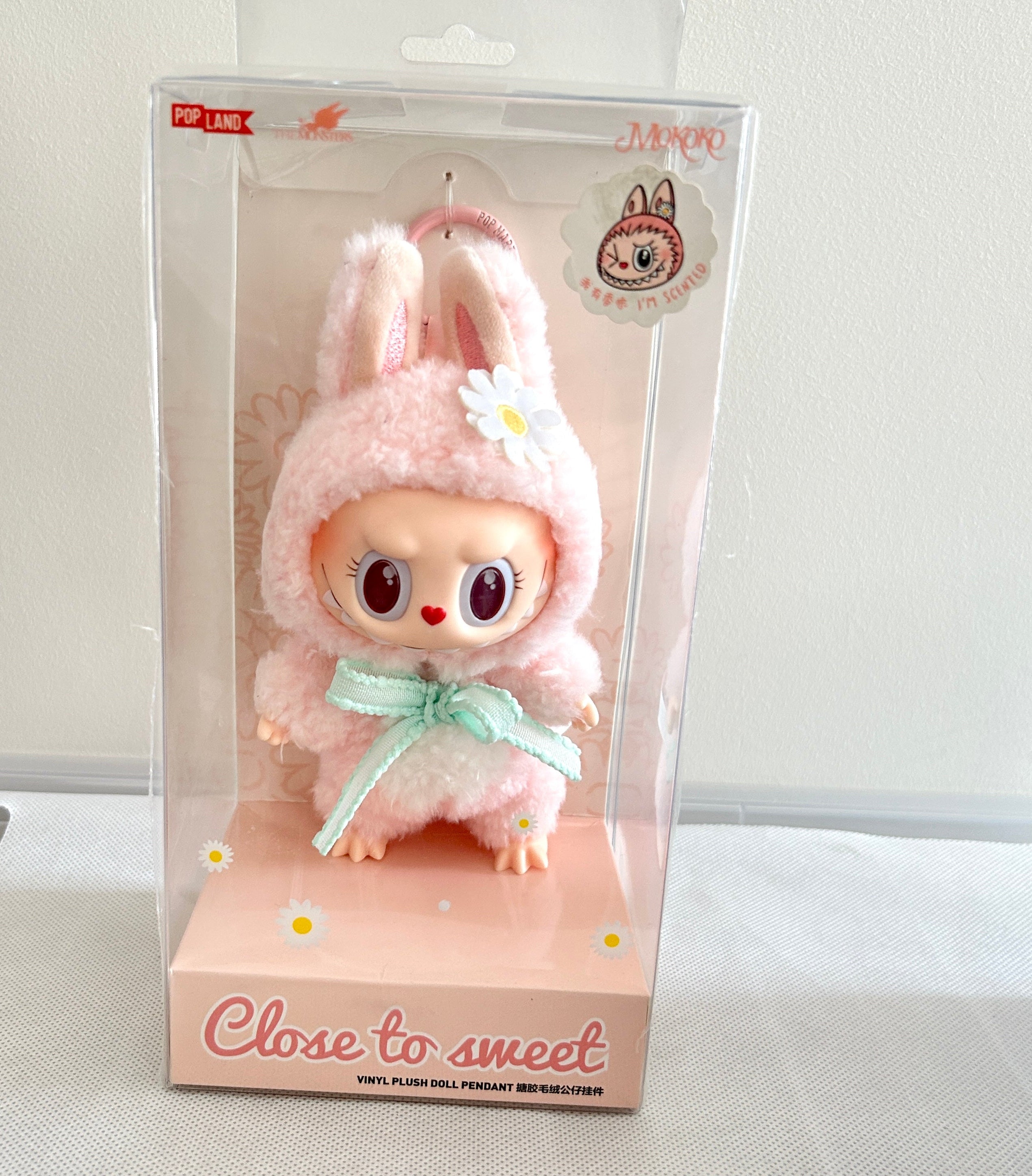 A plush doll character in a pink bunny outfit with a mint green bow, displayed inside a clear plastic retail box on a pink base. The box has 'Close to sweet' printed on the front.