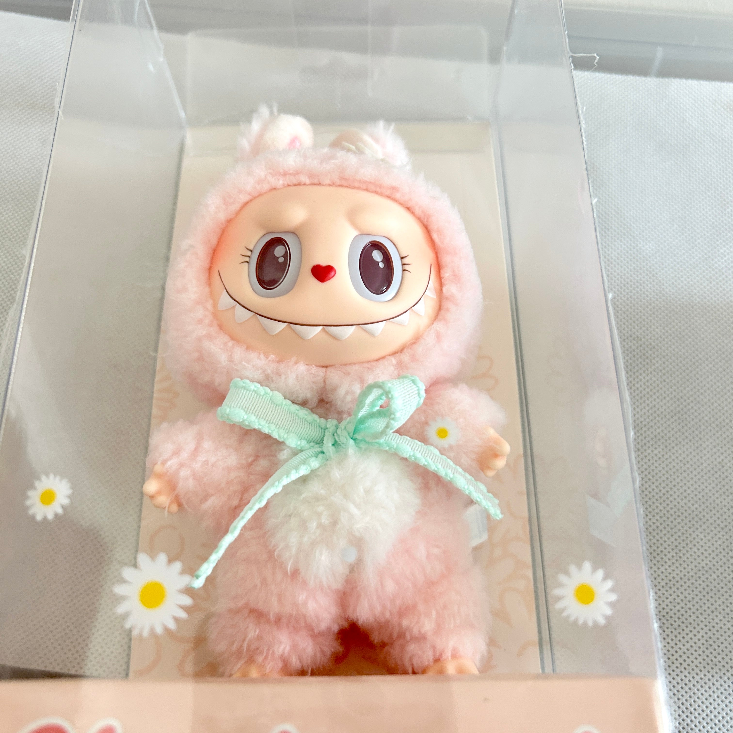 A plush toy doll in a pink fuzzy hooded outfit with a mint green bow, displayed inside a clear plastic box with daisy graphics on the front.
