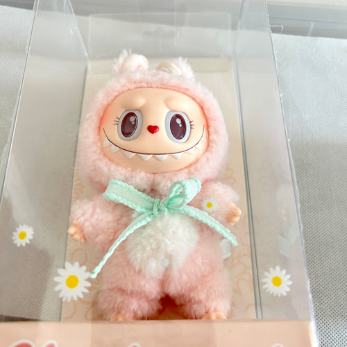 A plush toy doll in a pink fuzzy hooded outfit with a mint green bow, displayed inside a clear plastic box with daisy graphics on the front.