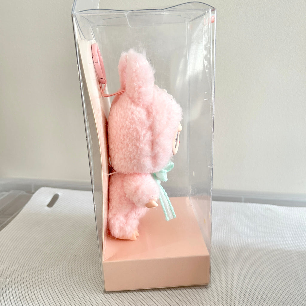 A pink fluffy plush toy character inside a clear plastic display box with a peach-colored base.