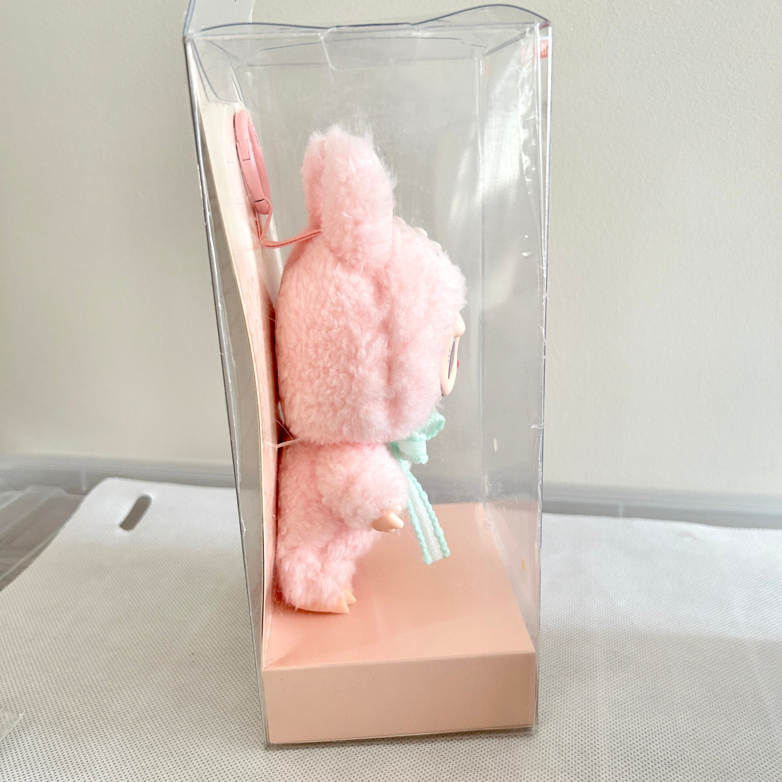 A pink fluffy plush toy character inside a clear plastic display box with a peach-colored base.