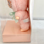 A small plush doll with fluffy pink fur, peach-colored limbs, and a mint green ribbon bow, displayed inside a clear plastic box with a pale pink base.