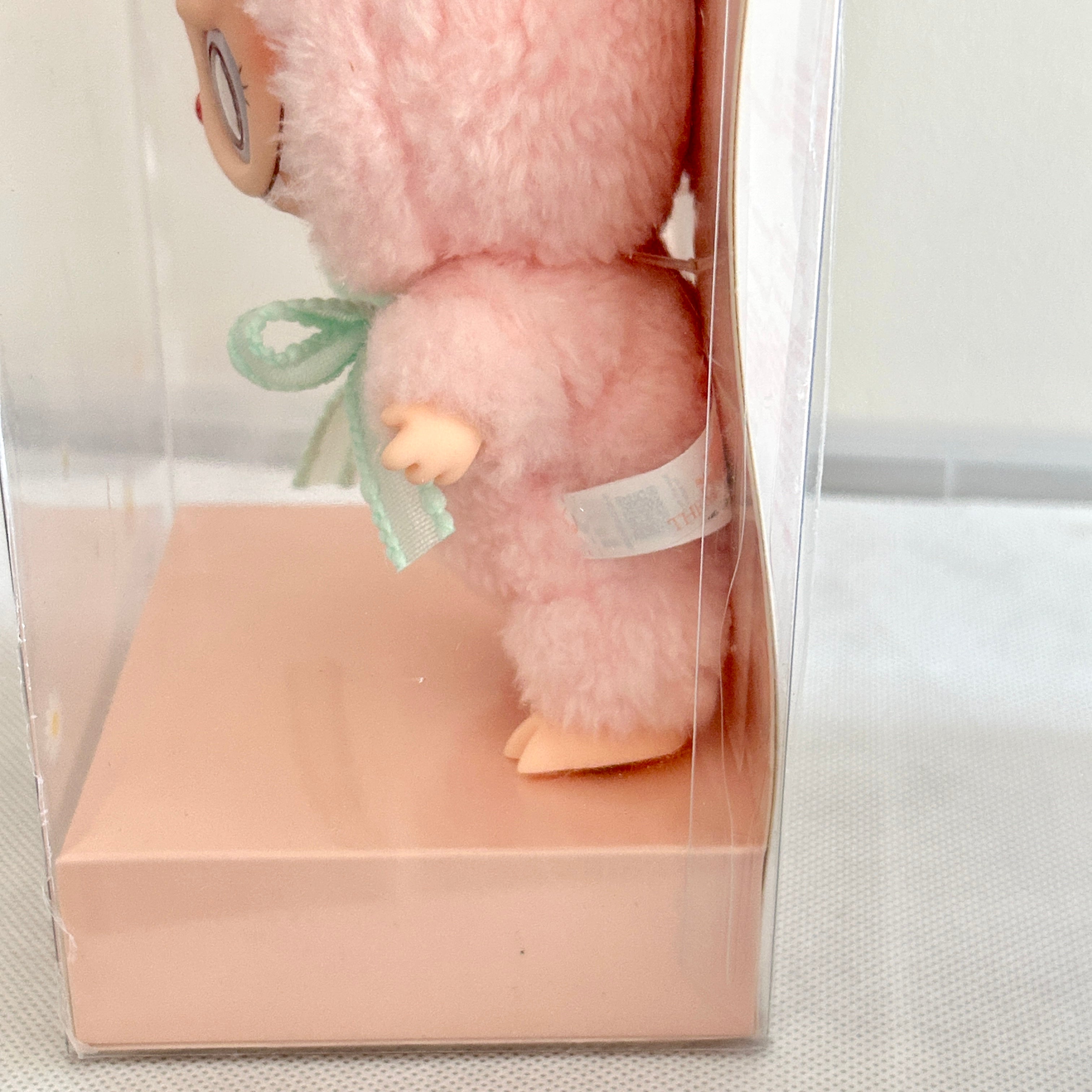 A small plush doll with fluffy pink fur, peach-colored limbs, and a mint green ribbon bow, displayed inside a clear plastic box with a pale pink base.
