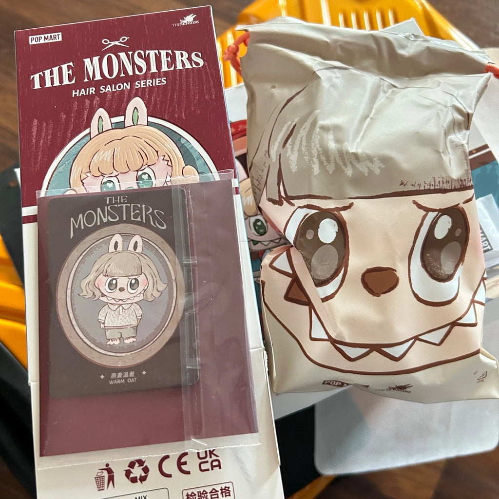 A collectible pin from 'The Monsters Hair Salon Series' by Pop Mart, shown next to its original maroon packaging box and a matching beige blind bag featuring a cartoon monster face.