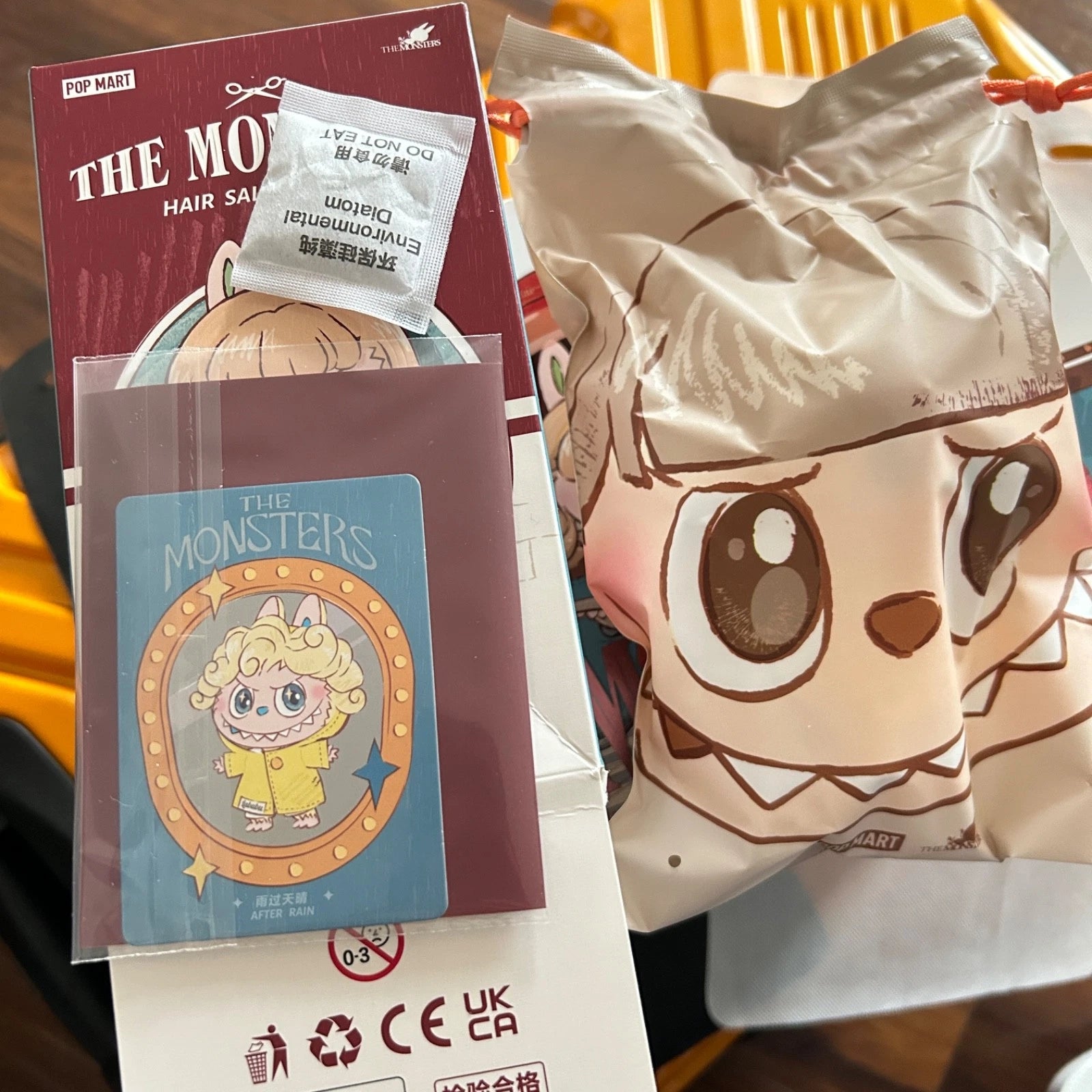 A mystery blind bag product set from Pop Mart's 'The Monsters' collection, featuring a sealed character bag with a cartoon monster face, a blue character card, and a hair spray packet on a white surface.