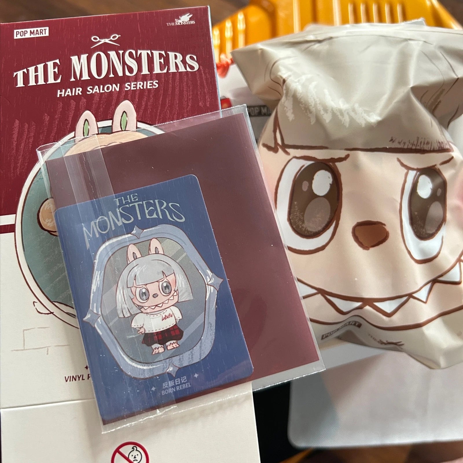 A collectible card from The Monsters Hair Salon Series by Pop Mart, sealed in a protective sleeve and displayed next to its red packaging box and a large character-themed blind bag.