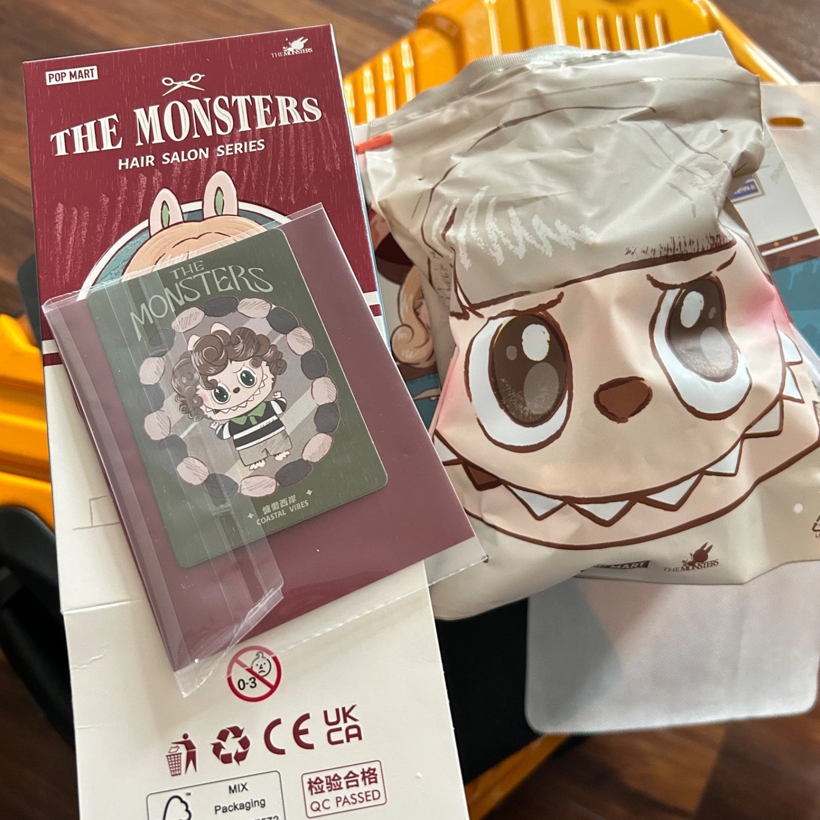 A close-up of a character-themed hair salon set featuring a brown plush toy with a cartoon face, a green hair clip with a cartoon girl illustration, and a red book or card titled 'THE MONSTERS HAIR SALON SERIES'.