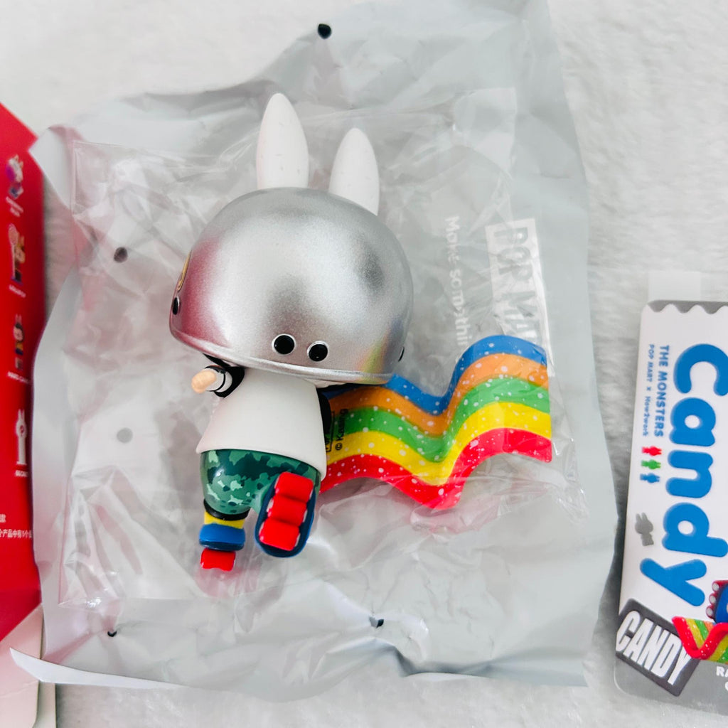 A collectible toy figure of a white rabbit with a metallic silver helmet and a rainbow cape, shown inside its clear plastic packaging next to a red box and a Candy: The Monsters card.