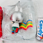 A collectible toy figure of a white rabbit with a metallic silver helmet and a rainbow cape, shown inside its clear plastic packaging next to a red box and a Candy: The Monsters card.