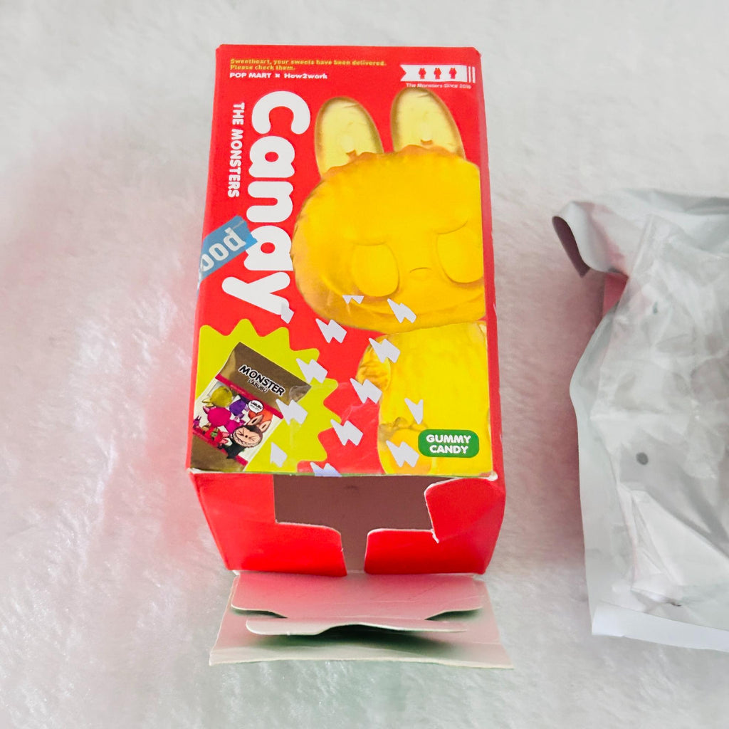 A red and yellow box of Pop Mart The Monsters Candy, featuring a large yellow monster graphic on the front, shown next to a silver foil packet on a textured white surface.