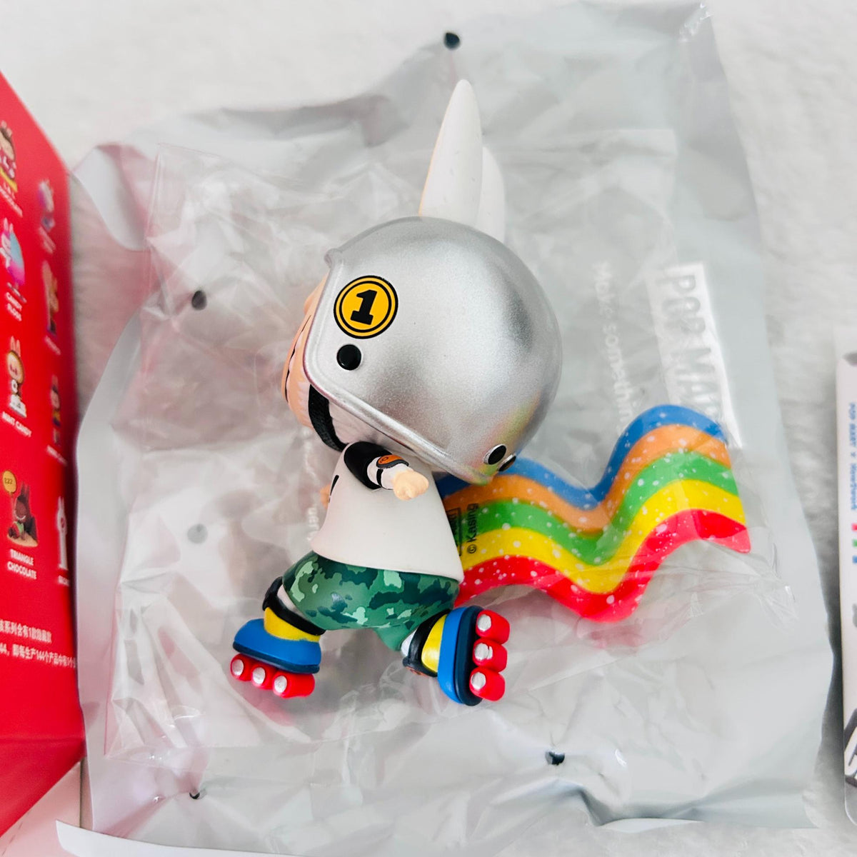 A collectible vinyl figure of a cartoon character wearing a metallic silver helmet and a green camouflage outfit, with a wavy rainbow-colored tail. The figure is shown inside its clear plastic blister packaging, with a red box partially visible on the left.