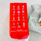A red collectible blind box showing illustrated character labels on the side, photographed on a light surface with part of a white shipping bag visible at the right.