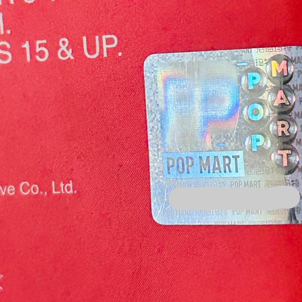 A close-up of a holographic POP MART sticker applied to a red textured surface, with part of the brand name 'POP MART' visible on the sticker and additional white text on the red background.