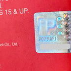 A close-up of a holographic POP MART sticker applied to a red textured surface, with part of the brand name 'POP MART' visible on the sticker and additional white text on the red background.
