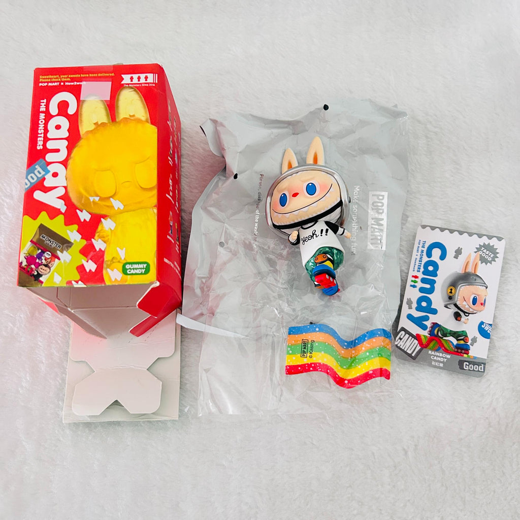 A collectible toy figure labeled 'Candy' shown with its red retail box, a clear plastic bag, and a small back card on a white textured surface. The figure has bunny ears, a white space-like helmet, and colorful rainbow accents.