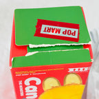 A close-up of a red and green Pop Mart candy box with a torn red sticker on top showing the brand name 'POP MART' in reverse, and a yellow wrapper partially visible at the bottom.