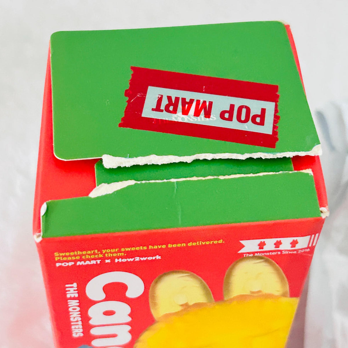 A close-up of a red and green Pop Mart candy box with a torn red sticker on top showing the brand name 'POP MART' in reverse, and a yellow wrapper partially visible at the bottom.