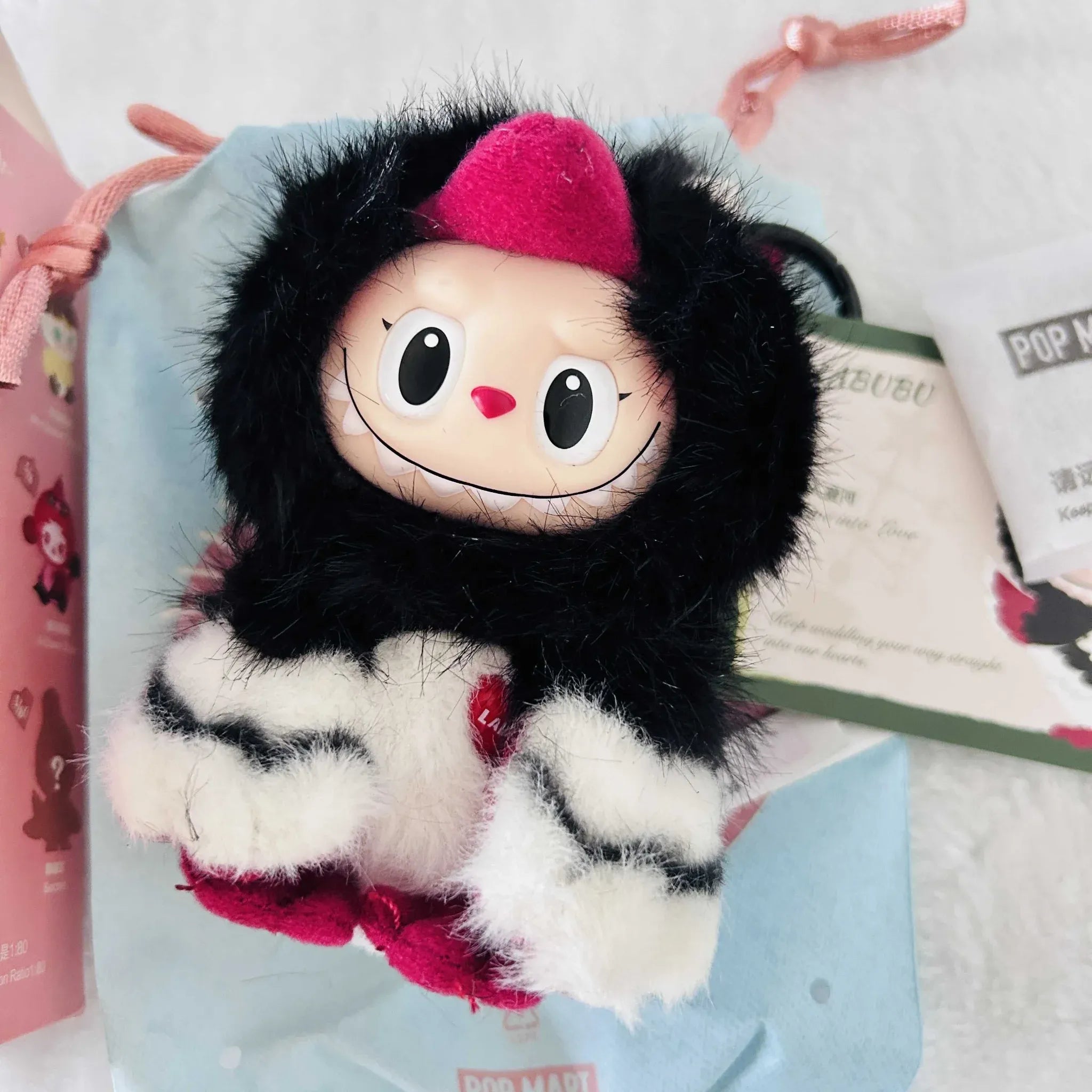 A Labubu Dear Birds Series plush toy, shows a black furry hood, a smiling beige face, resting on a  pouch with POP MART branding.