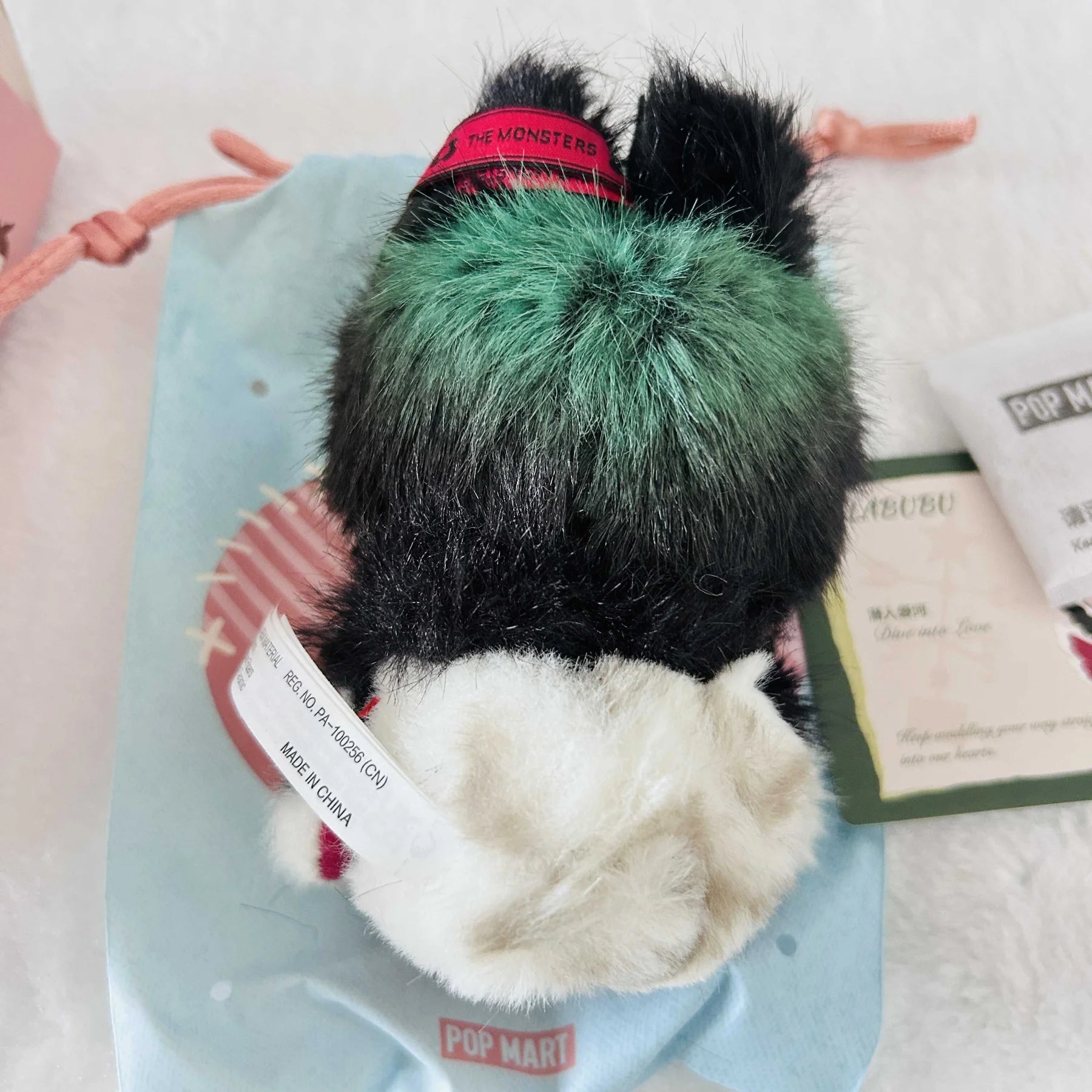 A back view of a Labubu with black, green, and white faux fur, shown from the back on a light blue POP MART pouch.