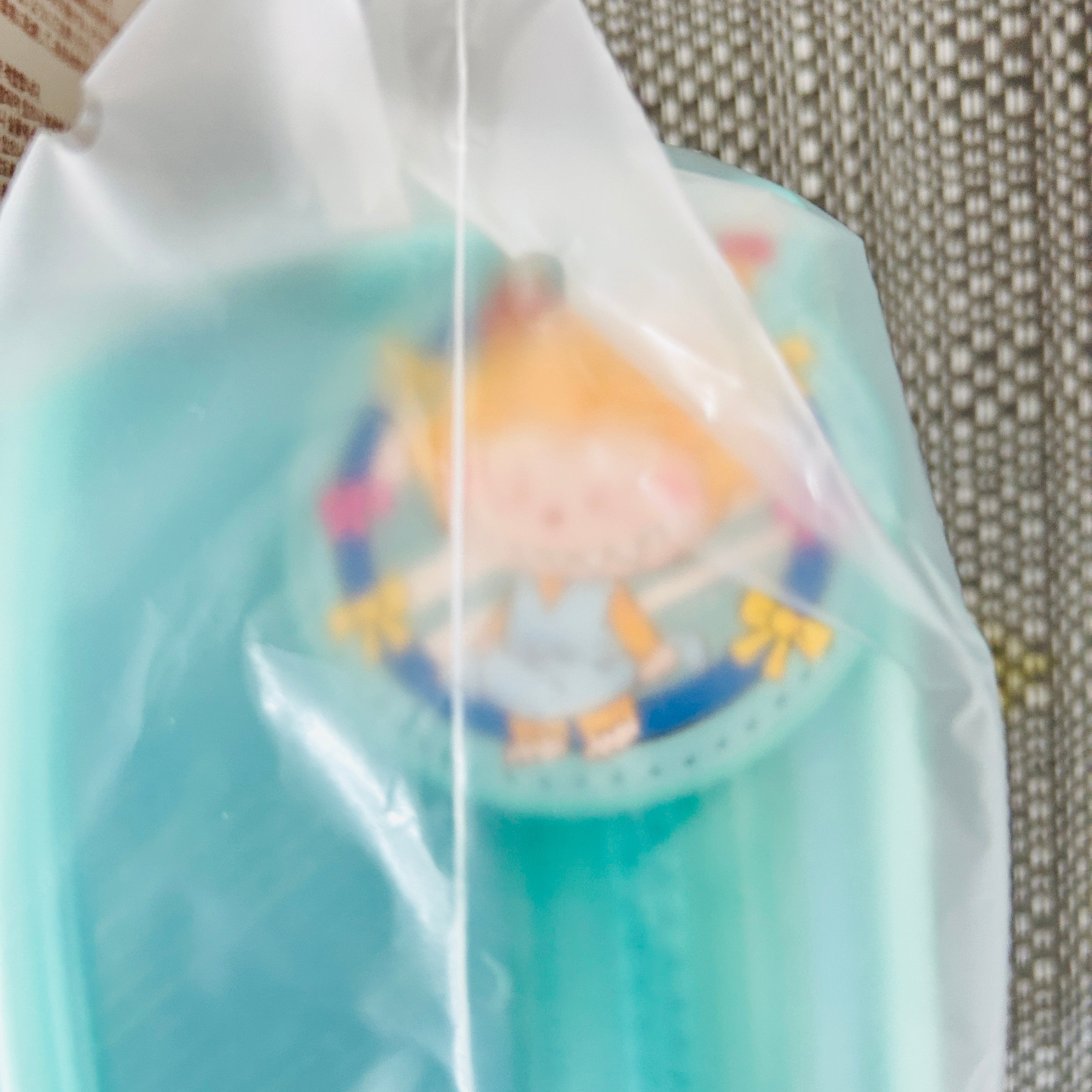 A light blue plastic bag containing a small round character accessory, photographed close up against a textured gray fabric background.
