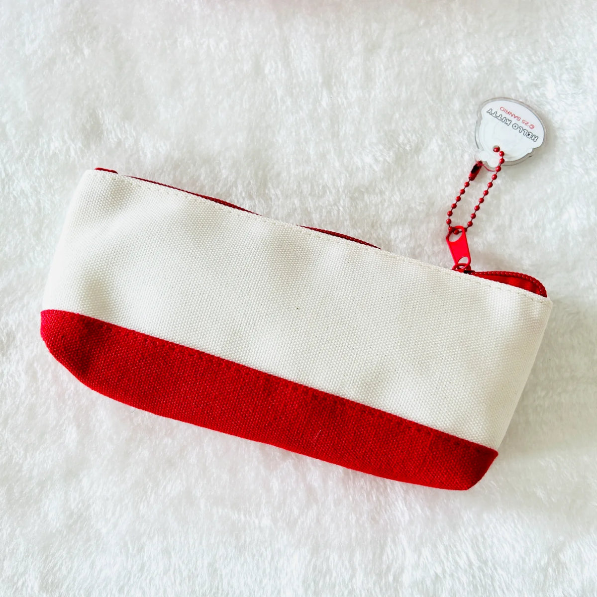A small zippered pouch with a cream-white top half and a red bottom half, photographed on a white textured faux-fur surface. A small attached hangtag is visible near the zipper.